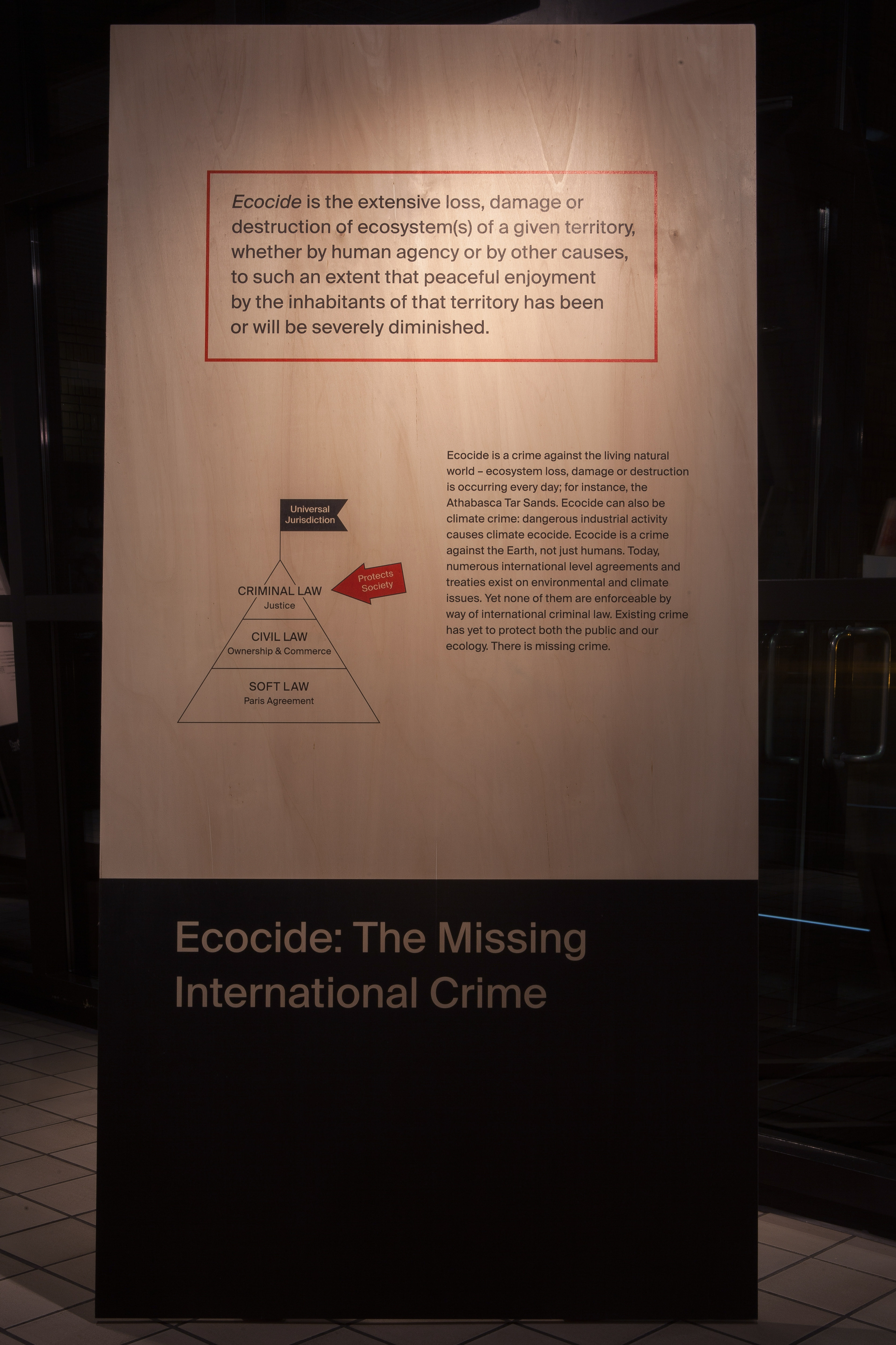 Installation. A floating wall panel is printed with the definition of ecocide, a pyramid graph with additional text, and the title below, which reads “Ecocide: The Missing International Crime”.