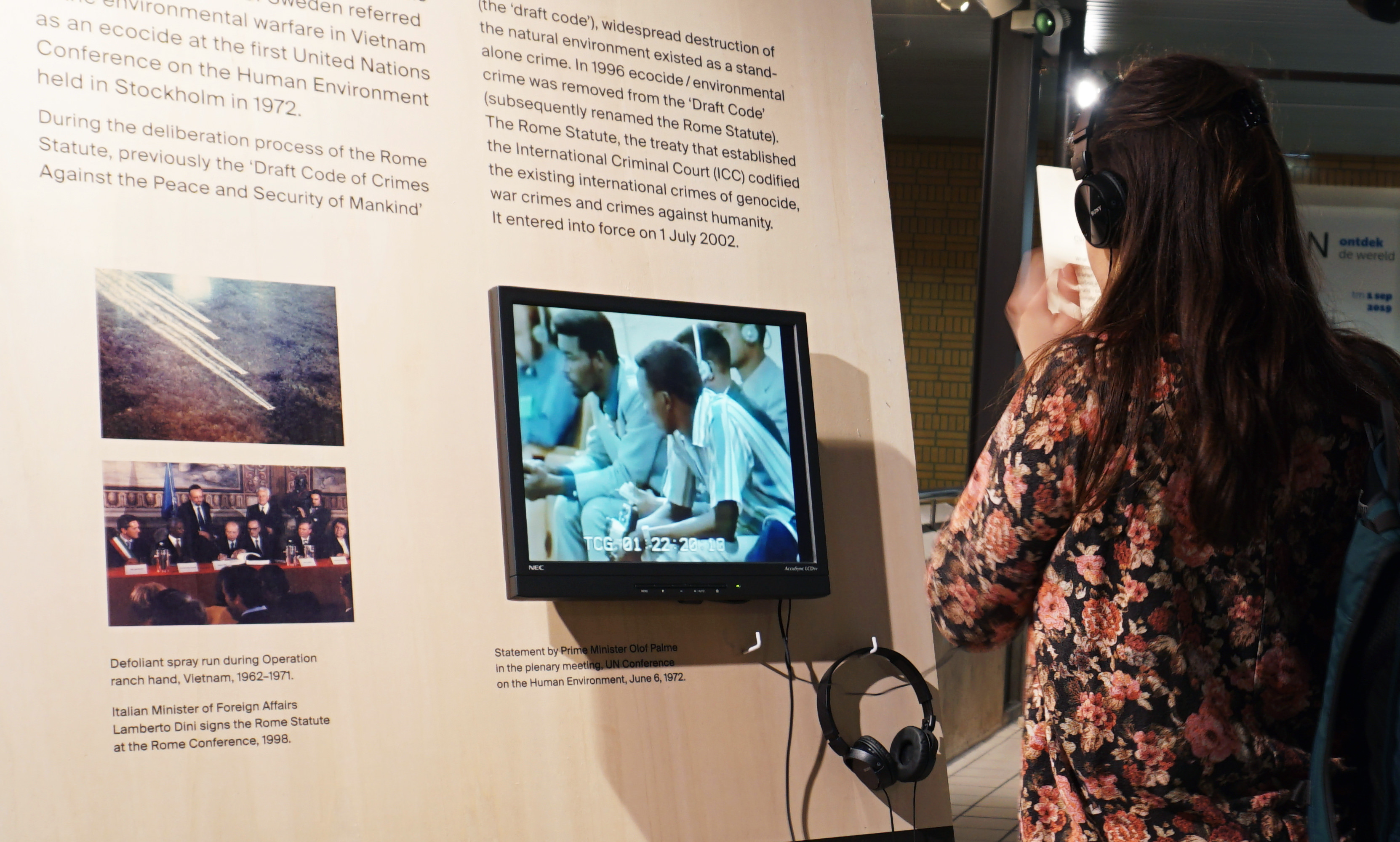 Installation. A woman listens to audio accompanying a video screen on a wall panel. Two colour photographs with captions are two our left and text is above.