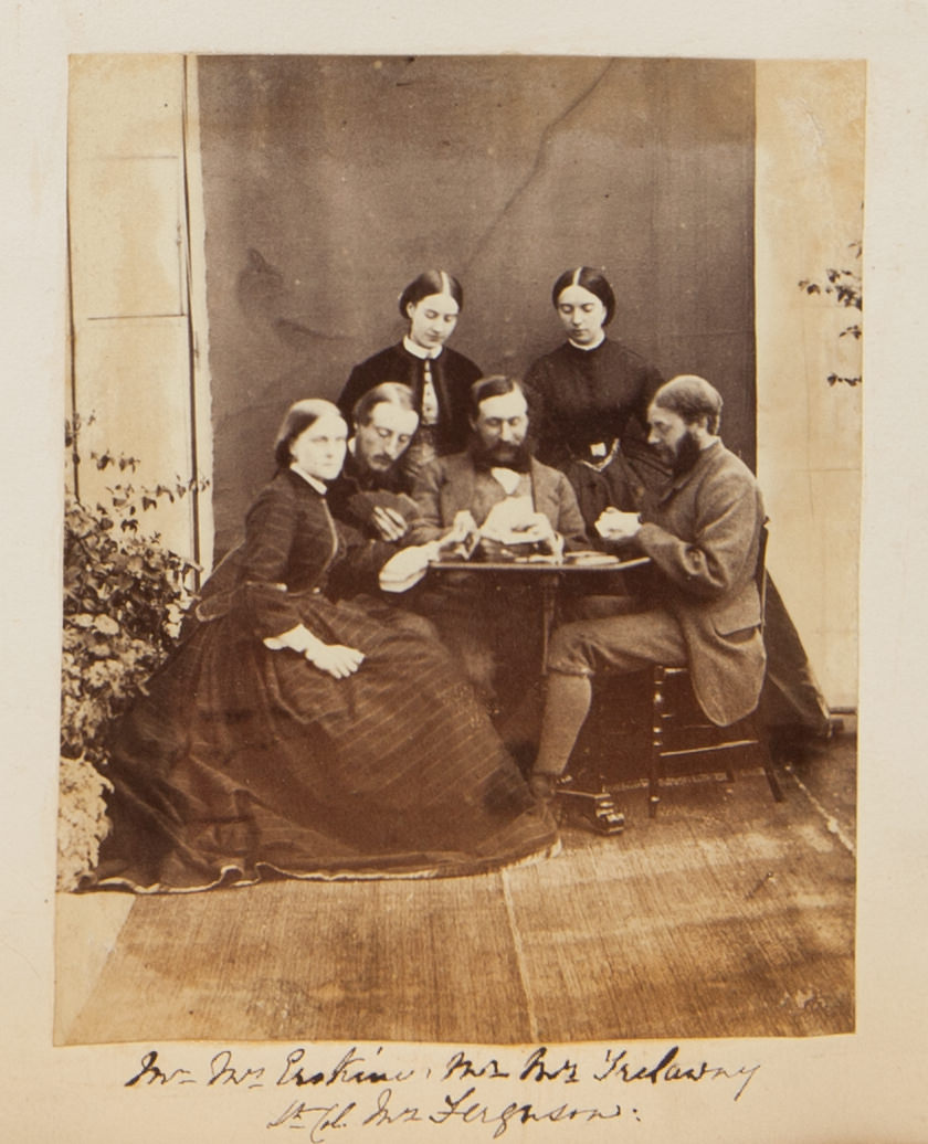 Black and white photograph. Three men and one woman sit playing cards at the far side of a small gaming table. Two more women stand behind the men and look down. All the people have pale skin.