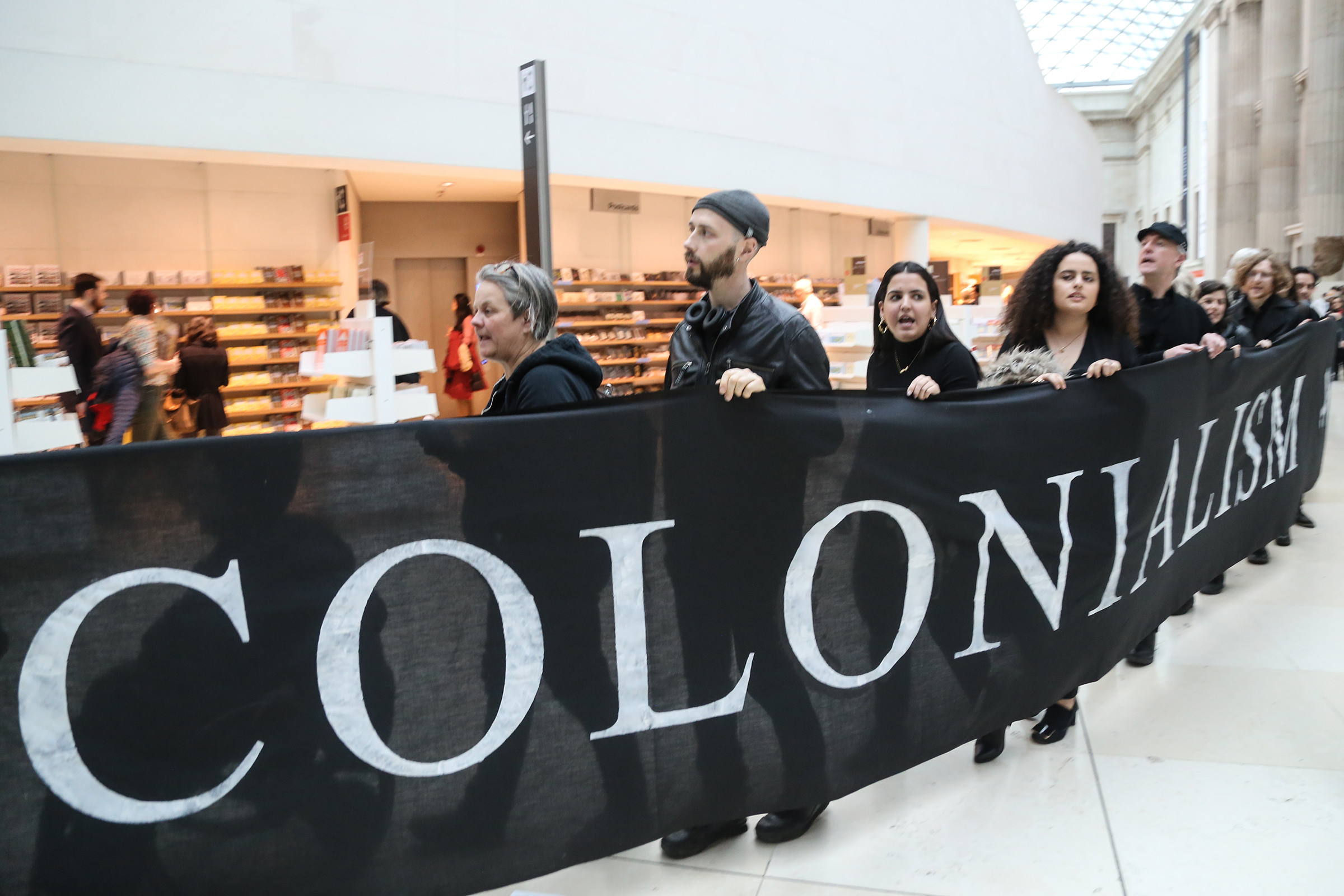 About a dozen people hold a black banner with white lettering reading “Colonialism”. The banner comes up to almost their shoulders, and it stretches in front of a gift shop.