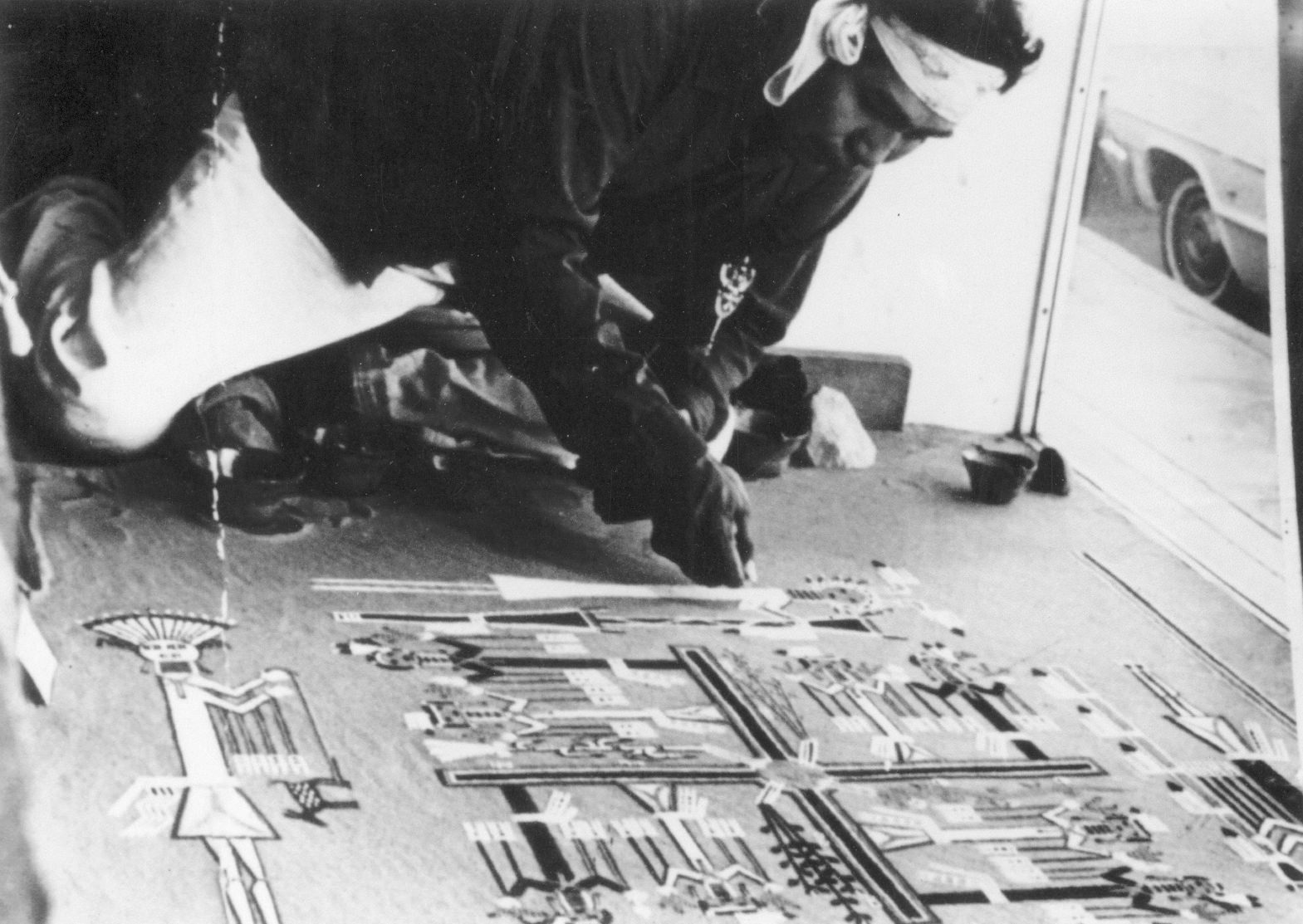 Installation. A dark-skinned man carefully drops sand from his fist to create a stylized image of people and natural forms like trees. He works on the floor, and the back end of a car can be seen out a window next to the artist.