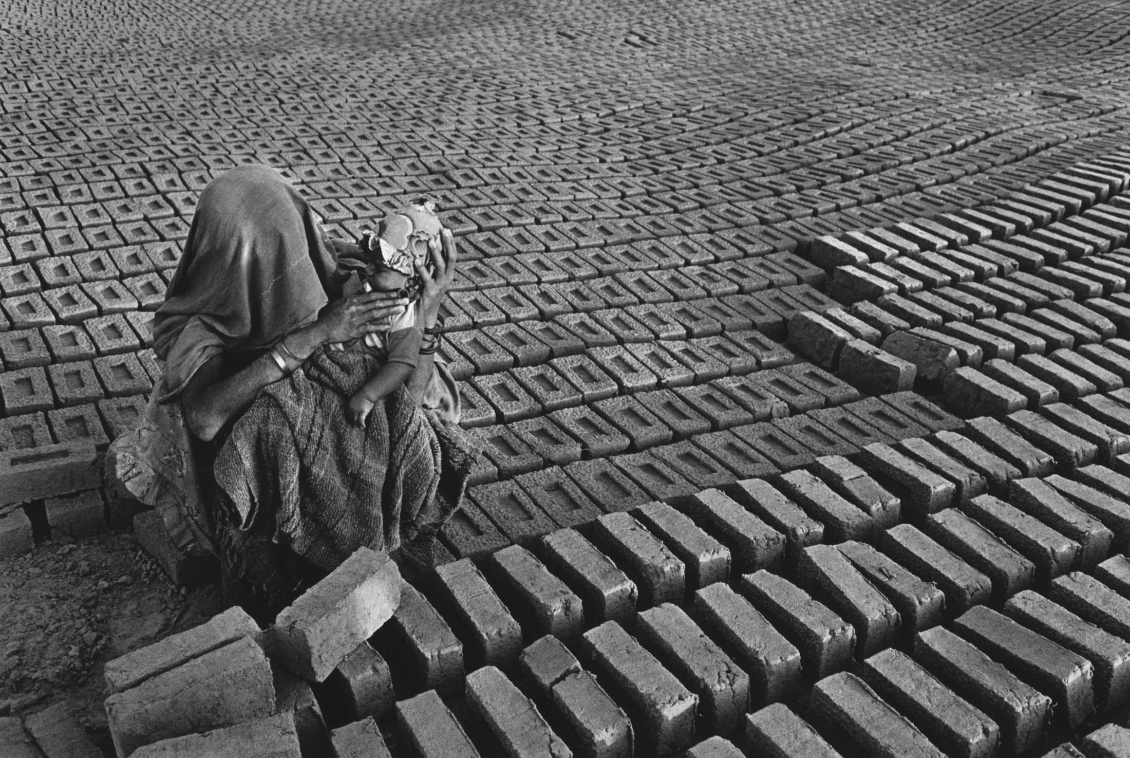 Black and white photograph. We look slightly down onto a woman sitting and holding a baby amid rows of bricks that surge and ebb with the lay of the land. The woman and baby’s faces and most of their bodies are covered with patterned cloth but their arms are dark skinned.